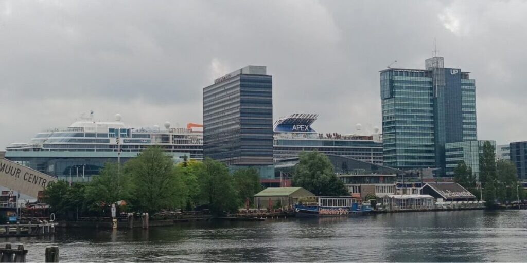 The Celebrity Apex cruise ship docked in a city port, surrounded by modern office buildings and lush greenery. The ship's name is visible on the upper decks, and the area features a mix of urban and natural elements, with a body of water in the foreground reflecting the cloudy sky.