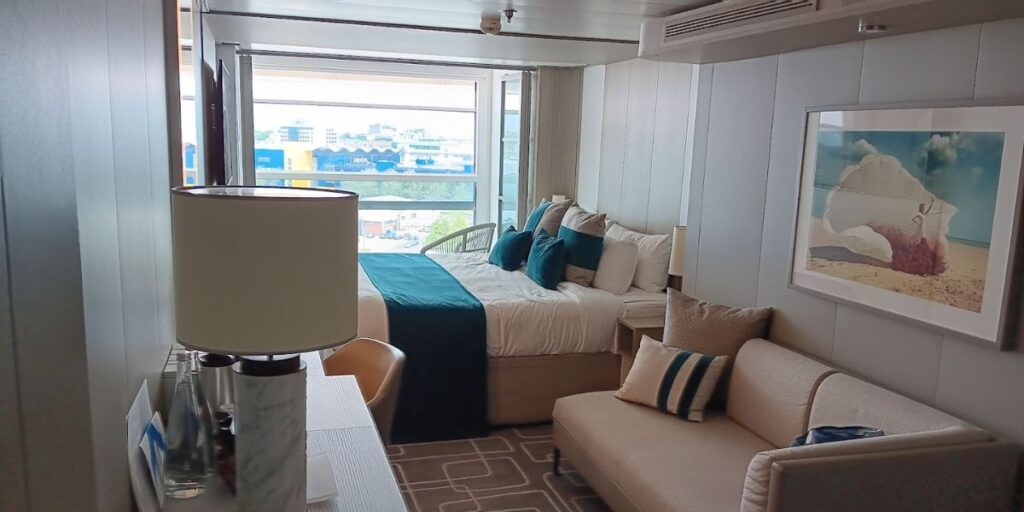 A modern stateroom on the Celebrity Apex cruise ship, featuring a neatly made bed with teal accents and an adjacent seating area with a sofa and pillows. The room has a large window providing a view of the cityscape, including an IKEA store, and is decorated with a beach-themed artwork.