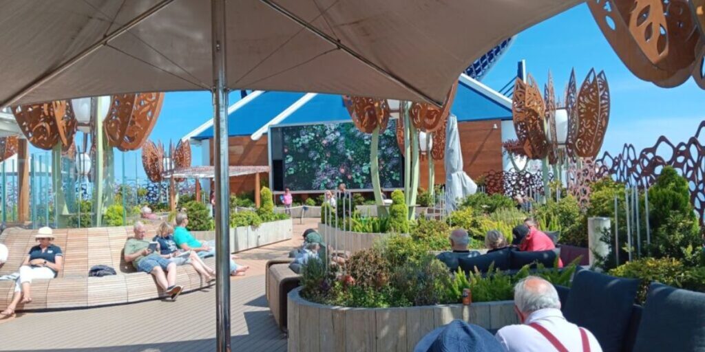 Passengers relaxing in the Rooftop Garden on the Celebrity Apex cruise ship, enjoying the sunny weather. The area features comfortable seating, lush greenery, and decorative metal sculptures, with a large screen displaying a floral image in the background.
