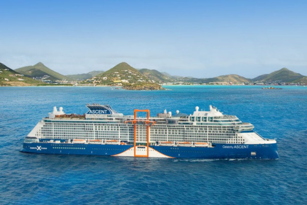 This image captures the Celebrity Ascent cruise ship sailing through bright blue waters with lush green islands in the background. The ship's sleek design and expansive deck space are complemented by the distinctive orange feature known as the "Magic Carpet." The clear, sunny weather and calm seas enhance the scenic tropical setting, perfect for a luxury ocean voyage.