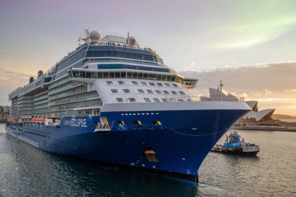 This image shows the Celebrity Eclipse cruise ship arriving in Sydney, with the iconic Sydney Opera House visible in the background. The ship's sleek, modern design stands out as it glides through the harbor at sunset, creating a beautiful contrast between the deep blue hull and the warm hues of the evening sky. A tugboat can be seen assisting the ship as it makes its approach to the dock, emphasizing the grandeur of the vessel against the scenic city backdrop.