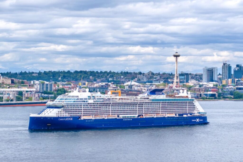This image shows the Celebrity Edge cruise ship sailing near a cityscape with the iconic Space Needle prominently visible in the background. The ship's modern design, including multiple decks and a sleek blue hull, stands out as it glides through the water. The cloudy sky and urban skyline create a scenic contrast with the ship, highlighting the blend of luxury cruising and metropolitan views.