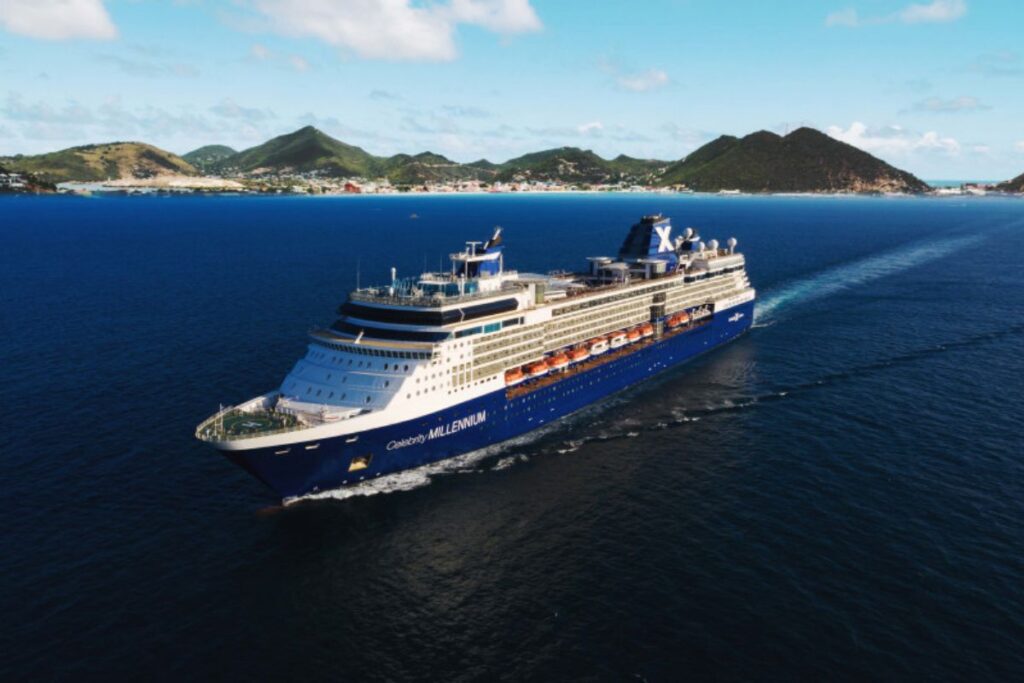 This image features the Celebrity Millennium cruise ship sailing through the calm blue waters with a picturesque island in the background. The ship’s distinctive blue and white exterior stands out against the ocean and the lush, green hills of the island. The bright, sunny day and clear skies emphasize the serene, tropical setting as the ship leaves a smooth wake behind it.