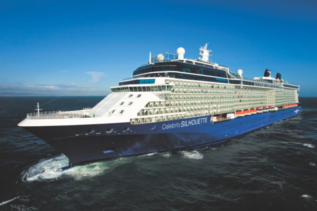 This image features the Celebrity Silhouette cruise ship sailing through open waters under a clear blue sky. The ship’s sleek design includes rows of stateroom balconies, its distinctive white and blue hull, and spacious decks. The vessel is cutting through the calm ocean, leaving a trail of wake behind it, showcasing the grandeur and elegance of this luxury cruise liner.