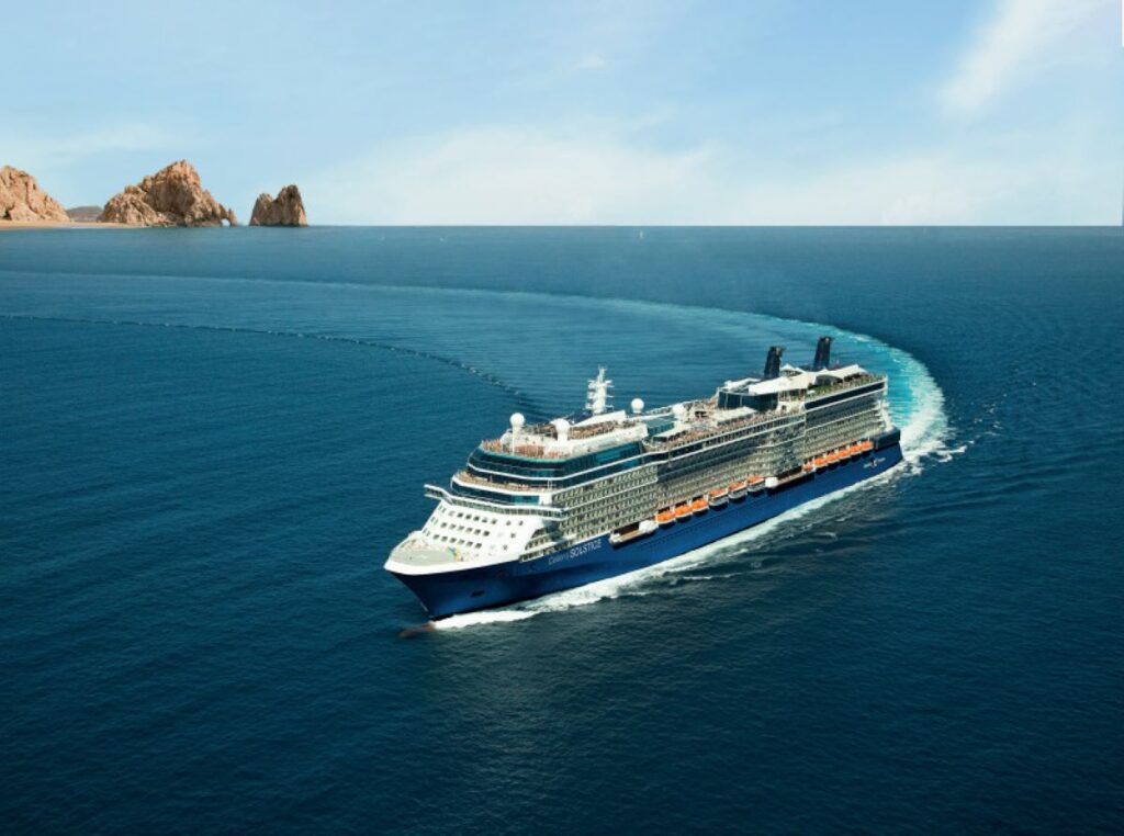 This image features the Celebrity Solstice cruise ship sailing through a calm, blue ocean with the famous rock formations of Cabo San Lucas visible in the distance. The ship is making a gentle turn, leaving a graceful wake behind it. The expansive sea and bright sky create a serene and picturesque backdrop, highlighting the luxurious vessel as it glides toward its destination.