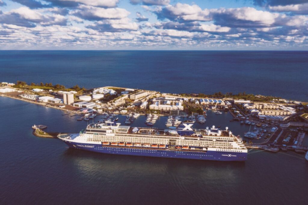 This image shows the Celebrity Summit cruise ship docked at a picturesque marina, with a sprawling waterfront complex and a calm, expansive ocean in the background. The ship’s bold design, featuring the prominent "X" logo on its funnel, contrasts beautifully with the clear skies and soft clouds. The aerial perspective highlights the serene coastal setting, with the ship docked alongside smaller vessels in the port.