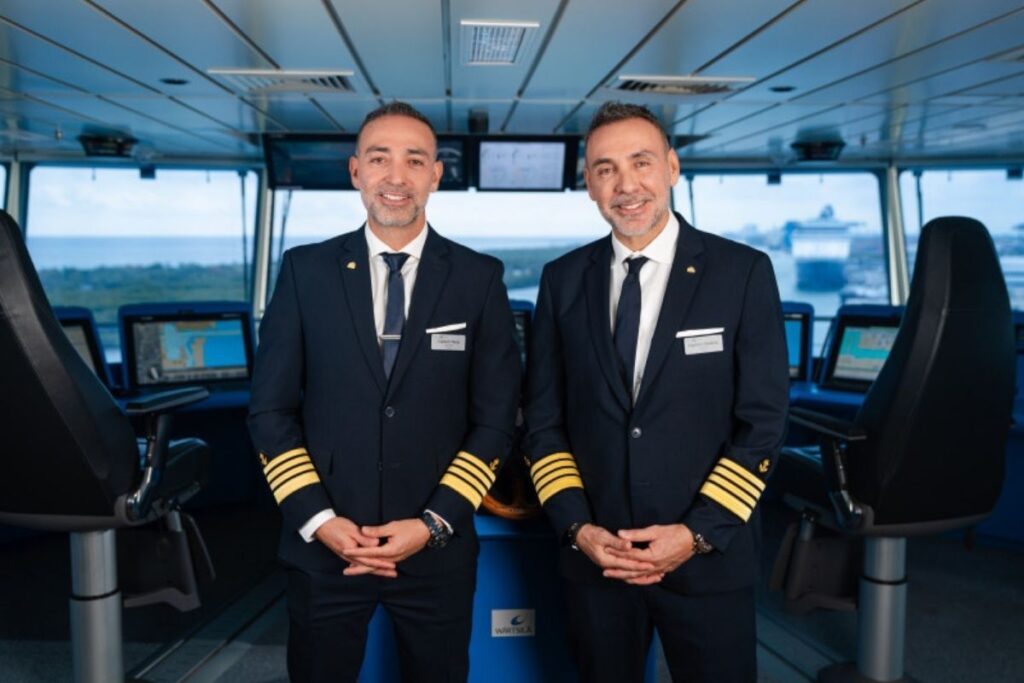 This image features two Celebrity Cruise ship captains standing on the ship's bridge, dressed in formal navy-blue uniforms with gold-striped epaulettes. Both captains are smiling confidently, with the modern navigation equipment and expansive sea visible in the background. The image highlights professionalism and leadership, emphasizing the expertise required to command a cruise vessel.