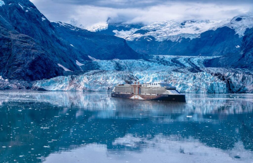 This image features a Celebrity cruise ship navigating through serene, icy waters surrounded by dramatic Alaskan landscapes. In the background, a stunning glacier cascades down into the water, framed by rugged snow-capped mountains under a cloudy sky. The ship's sleek design contrasts with the untouched natural beauty, emphasizing the unique experience of cruising through Alaska.