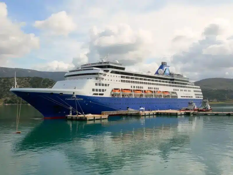 Large blue and white cruise ship named Celestyal Journey docked at a calm port with green hills and cloudy skies in the background. The ship is reflected in the still turquoise water, and lifeboats line its side above several passenger decks.