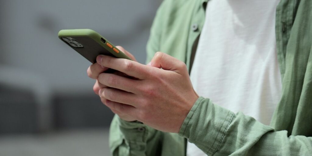 Close-up of a person texting on a cell phone, wearing a green overshirt and white T-shirt, with focus on their hands and the smartphone in a green case. The image captures a casual moment of cell phone texting in a modern, everyday setting.