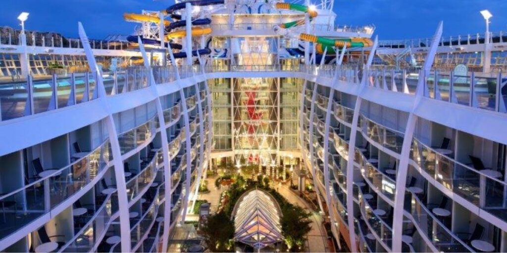 View of the Central Park neighborhood aboard Royal Caribbean’s Harmony of the Seas, showcasing lush greenery, walking paths, and restaurant seating nestled between towering balcony staterooms. Above, colorful waterslides stretch across the top deck under a twilight sky.
