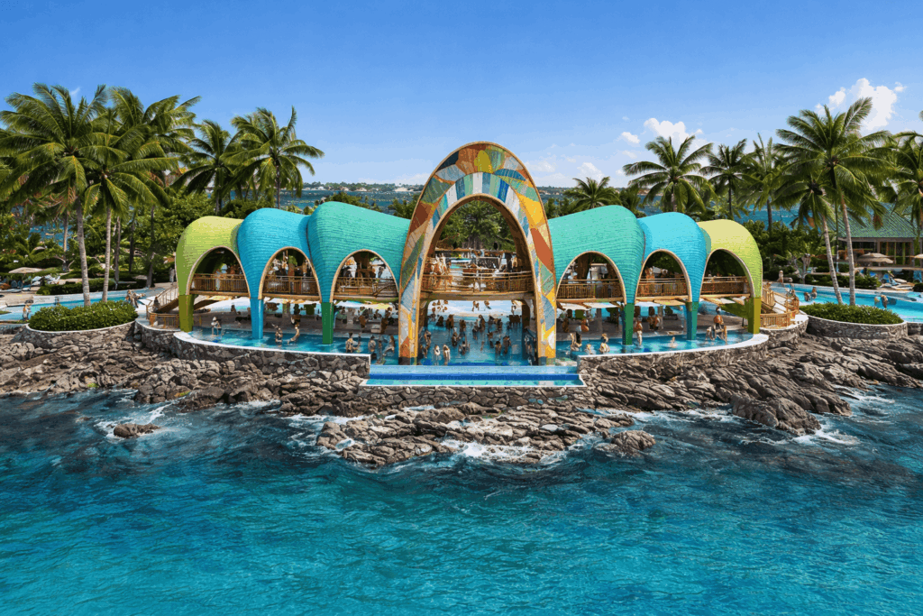 An aerial view of a colorful swim-up bar and pool complex built along a rocky shoreline, surrounded by turquoise ocean water and palm trees.