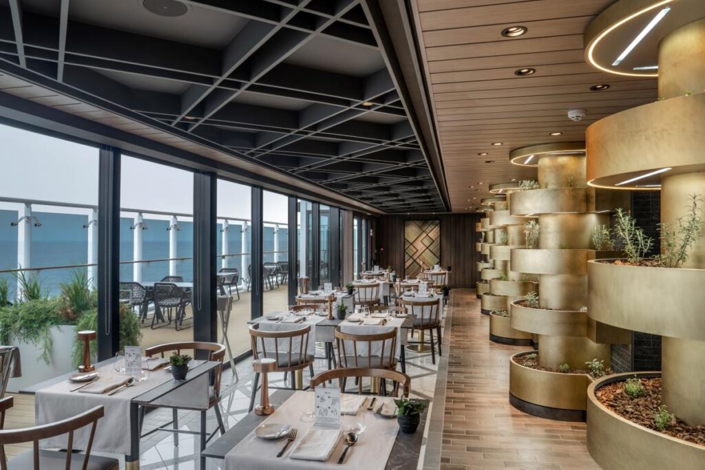 Chef's Garden Kitchen on MSC World Europa, featuring an elegant dining area with white tablecloths, wooden chairs, and a unique vertical garden with lit circular shelves, all alongside floor-to-ceiling windows offering ocean views.