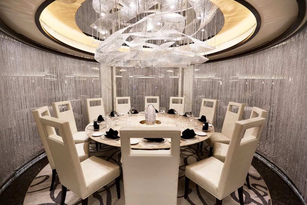 The image captures the opulent Chef’s Table Lumiere on the Majestic Princess cruise ship, designed for an exclusive dining experience. The elegantly set table with pristine white chairs is enclosed by a curtain of shimmering light strands, creating a sense of privacy and glamour. Above, an intricate chandelier adds a touch of luxury, and the warm lighting complements the sophisticated ambiance of this intimate culinary setting.