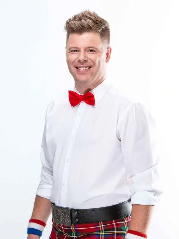Chris Williams, former Carnival cruise director known as the Flying Scotsman, smiling in a studio-style portrait wearing a white shirt, red bow tie, and tartan belt.