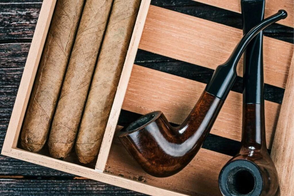 A wooden box filled with fine cigars adjacent to two elegant smoking pipes resting on a slatted wooden surface. The rich textures and warm tones of the wood convey a sense of traditional smoking luxury and the time-honored ritual of tobacco enjoyment.