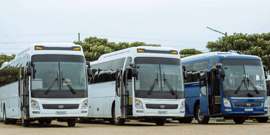 Three modern Hyundai tour buses parked side by side on a paved lot, with two in white and one in blue. The buses feature large tinted windows, destination displays, and visible Rwandan license plates, set against a backdrop of leafy trees and utility lines.