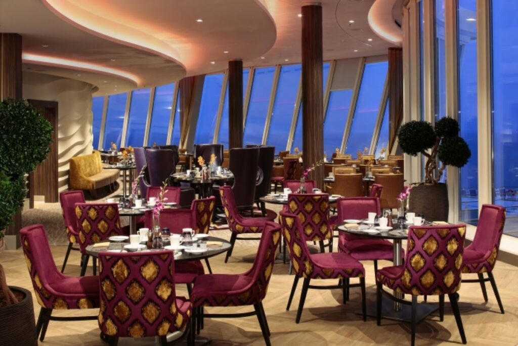 Elegant dining setup inside the Coastal Kitchen on Harmony of the Seas, featuring plush purple chairs, sophisticated table settings, and floor-to-ceiling windows that offer a panoramic view of the sea.