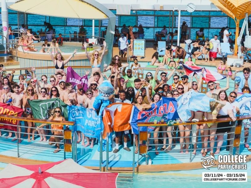A large group of young adults is gathered on the deck of a cruise ship for a college party event, many of them holding up flags representing different universities. The crowd, dressed in swimwear and sunglasses, is smiling, cheering, and posing for the camera while standing around a pool area. A mascot in a blue shark costume stands in the center, adding to the lively atmosphere. The "College Party Cruise" logo and event dates (March 11-15, 2024) are displayed in the bottom right corner of the image.