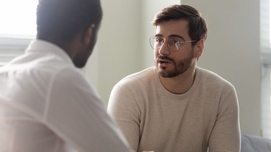 This image shows two individuals engaged in a serious conversation, with one person speaking while the other listens attentively. The setting appears casual and professional, suggesting a discussion or counseling session. 