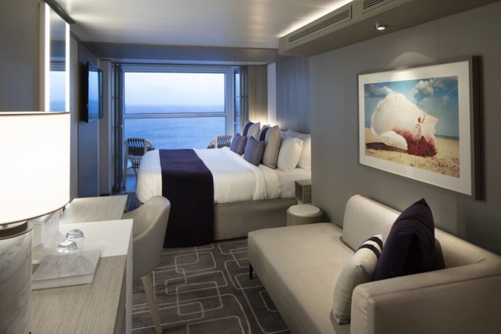 This image depicts a Concierge Class stateroom aboard a Celebrity cruise ship, showcasing its luxurious and modern design. The room features a plush bed with premium linens, a cozy seating area, and floor-to-ceiling windows with access to a private balcony for uninterrupted ocean views. Soft lighting, elegant artwork, and a neutral color palette with deep purple accents create a sophisticated and serene ambiance.