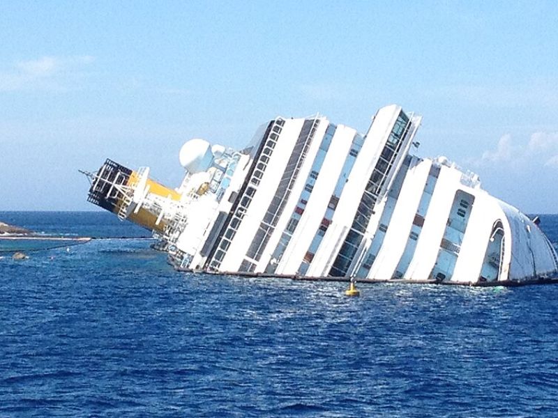 The Costa Concordia cruise ship is shown lying on its side in shallow blue water near the island of Giglio. The vessel is severely listing, with much of its white hull and yellow funnel partially submerged as it rests against the rocky coastline under a clear sky.