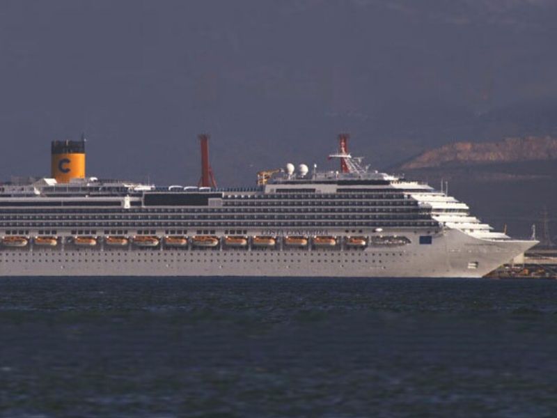 A large white cruise ship, the Costa Concordia, is shown sailing on calm water near the coastline. The vessel’s yellow funnel with a black “C” logo is visible, and passengers can be seen on the decks under a hazy sky with distant hills in the background.