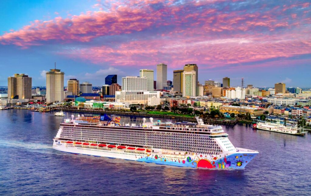 This image captures the Norwegian Breakaway cruise ship docked near the vibrant skyline of a city, with colorful, cloud-filled skies during sunset or sunrise. The ship's hull is adorned with lively, artistic designs, reflecting a fun and energetic vibe. The backdrop of tall buildings and a riverboat complements the modern ship, creating a striking blend of urban and maritime elements.