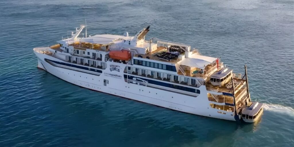 Aerial view of the Coral Adventurer cruise ship sailing on calm blue water, showing multiple decks, lifeboats, and outdoor seating areas. The ship was involved in the incident where an 80-year-old passenger was left behind on a remote island in Queensland, Australia.