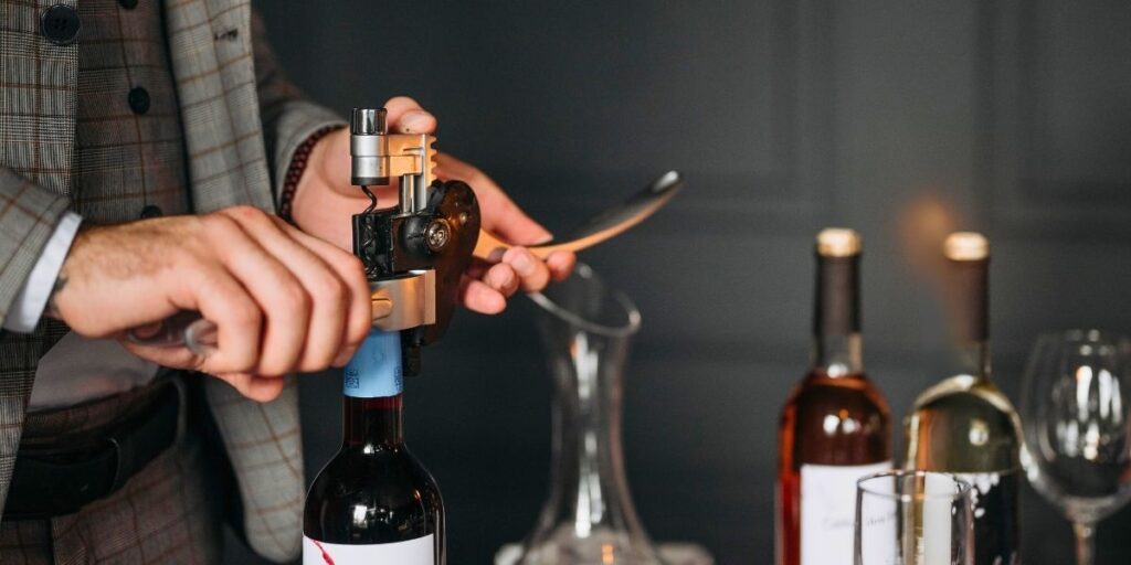 A person in a plaid suit uses a professional wine bottle opener to uncork a bottle of red wine, with additional wine bottles, glasses, and a decanter arranged on a dark tabletop. The scene suggests a refined, upscale wine service experience.