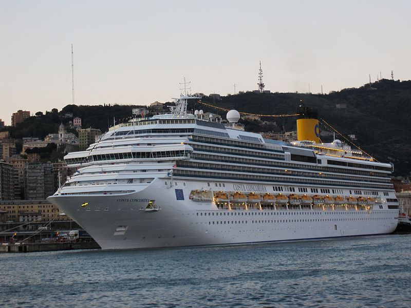 The Costa Concordia cruise ship is docked at port during early evening, its lights glowing along the decks. The large white vessel with a yellow funnel towers over the waterfront, with city buildings and hills rising in the background.