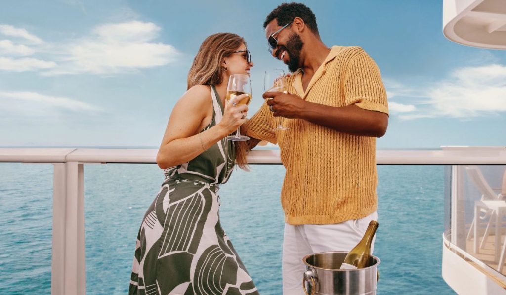 A cheerful couple enjoying champagne on a Carnival cruise balcony. The woman, wearing a stylish green and black patterned dress and sunglasses, toasts with the man clad in a summery yellow knit shirt and white pants. They share a moment of joy against a backdrop of calm blue seas and clear skies.