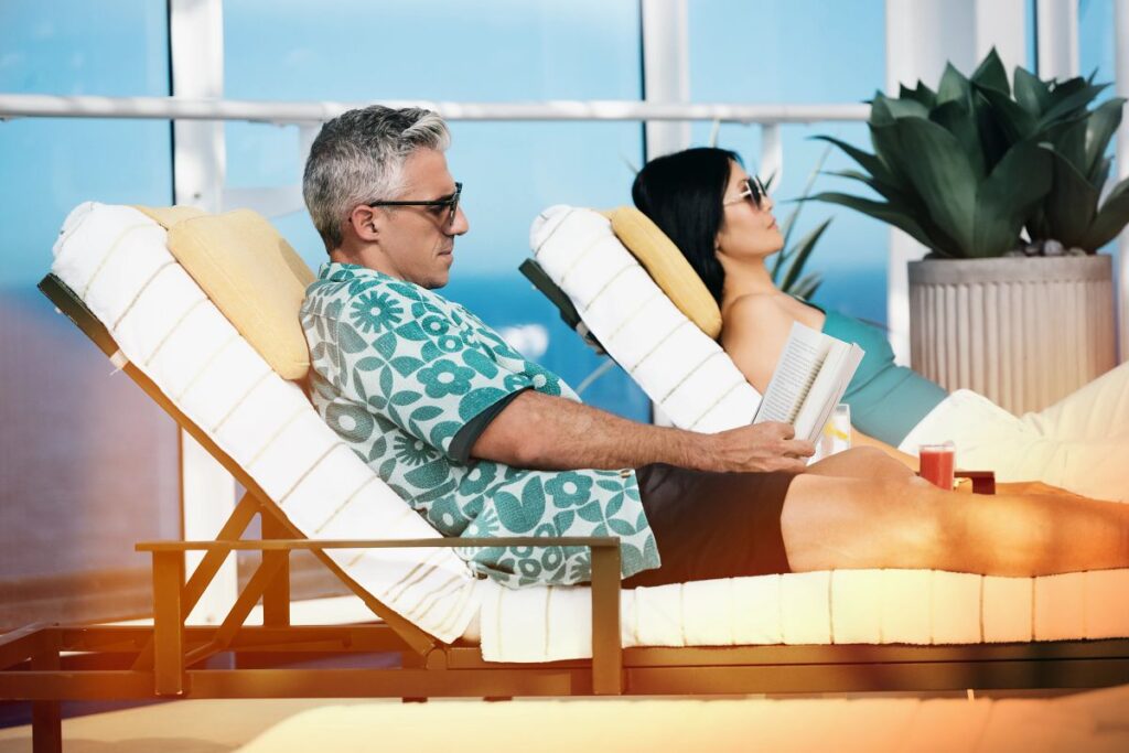 This image features a relaxed couple lounging on comfortable deck chairs aboard a Princess cruise ship. The man is reading a book, while the woman reclines with sunglasses, enjoying the serene atmosphere. With a backdrop of large windows showcasing ocean views, the scene exudes tranquility and leisurely luxury.