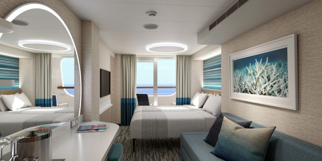 A modern Cove Balcony stateroom, bathed in natural light from the full-sized window offering a view of the sea. The room is tastefully decorated with a marine color palette, featuring a comfortable bed, a cozy sofa, and a contemporary design that includes a unique circular lighting element on the ceiling. A framed picture of coral on the wall accentuates the oceanic theme, enhancing the overall serene and stylish ambiance of the space.
