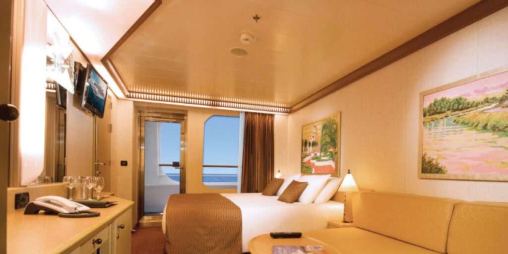 Elegant cove balcony cabin on a cruise ship, showcasing a comfortable queen-size bed with chocolate and cream bedding, a long cream sofa, and a warm-toned carpet. The room features a vanity area with a mirror, a flat-screen TV, and opens to a balcony with an ocean view, complemented by a vibrant landscape painting on the wall.