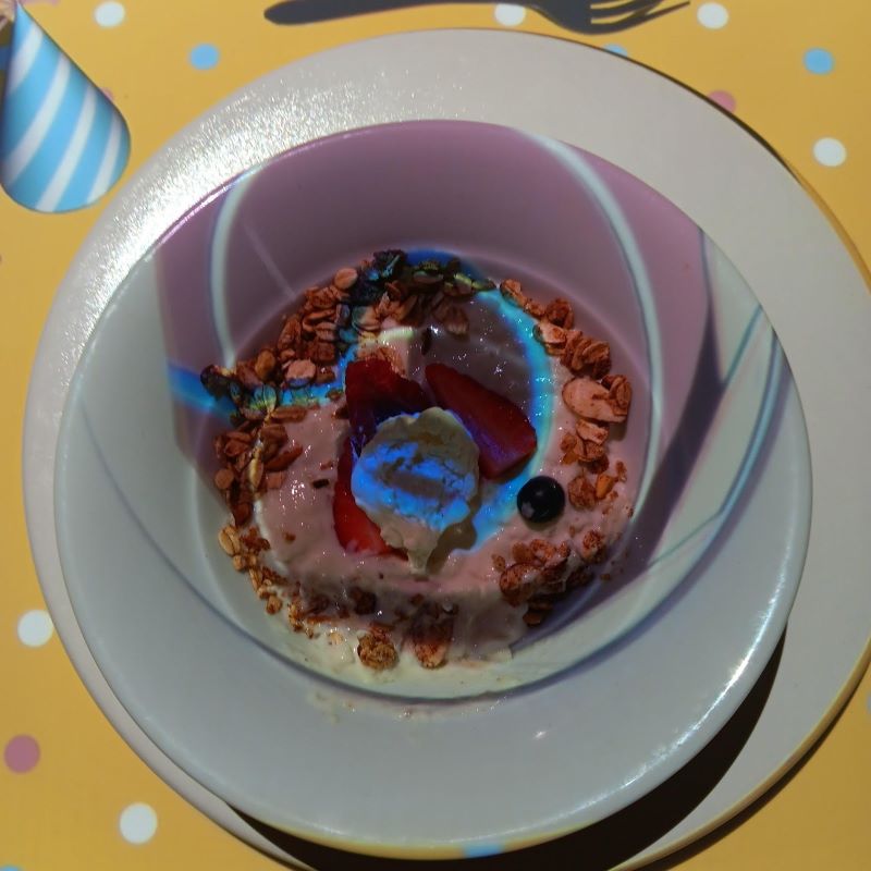 A bowl of creamy rice pudding is topped with fresh strawberries, blueberries, and a sprinkle of granola, garnished with a dollop of whipped cream. The dessert is set on a white plate with a colorful, playful projection on the table, adding a fun visual element to the dining experience at Le Petit Chef on the Celebrity Apex cruise ship.