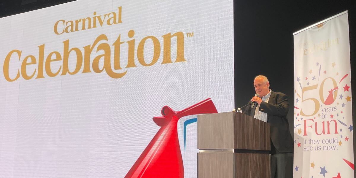 John Heald speaking at a podium during a Carnival Celebration event, with the Carnival Celebration logo displayed on a large screen behind him.
