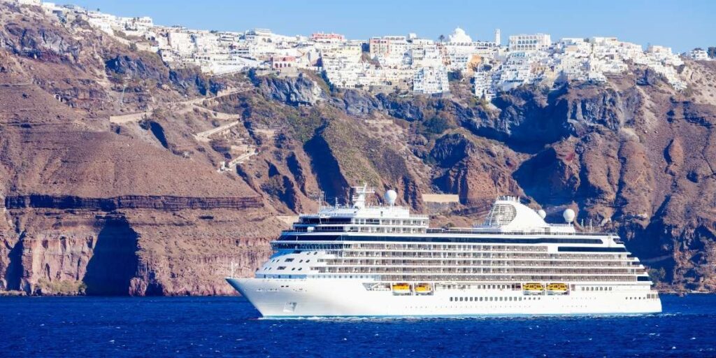 A large white cruise ship sails through the deep blue waters off the coast of Santorini, Greece, with the island's steep volcanic cliffs and iconic whitewashed buildings perched high above in the town of Fira. The dramatic contrast between the rugged brown terrain and the crisp architecture highlights the island’s unique beauty.
