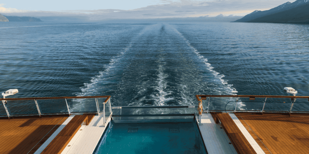 Cruise ship stern view with an infinity pool and teak deck in the foreground, looking out over the ship’s wake with mountains under a blue sky.