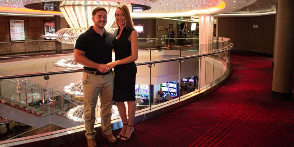 A couple embraces and smiles on the deck of a cruise ship at sunset, with the ocean stretching out behind them. The woman wears a light dress and heels, while the man is dressed in white, both glowing in the warm golden light.