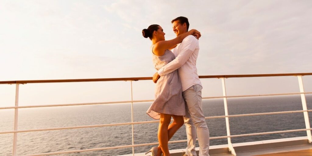 A couple embraces and smiles on the deck of a cruise ship at sunset, with the ocean stretching out behind them. The woman wears a light dress and heels, while the man is dressed in white, both glowing in the warm golden light.