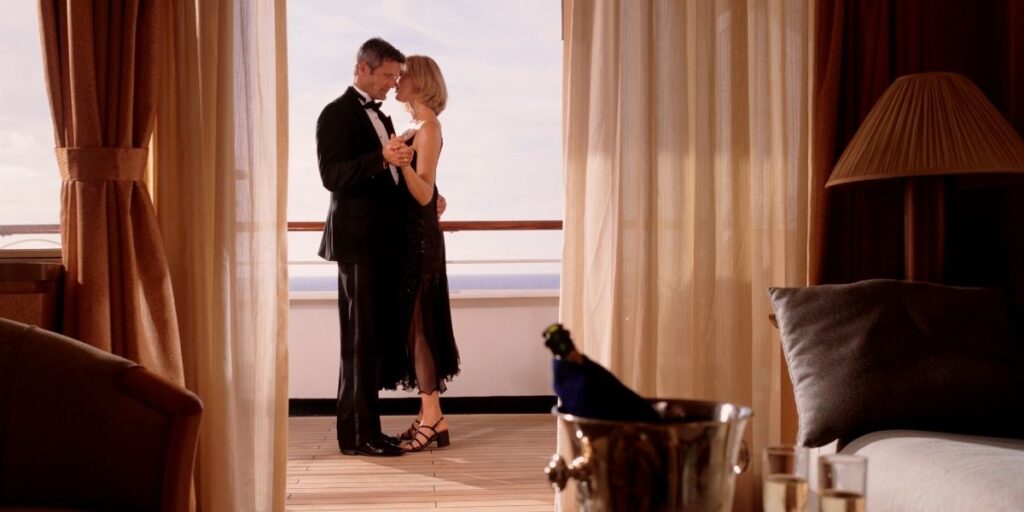 An elegantly dressed couple shares a quiet dance on the balcony of a cruise ship suite, framed by flowing curtains. The man wears a black tuxedo and the woman a black evening dress, with champagne and flutes set on a nearby table, creating a romantic and luxurious atmosphere.