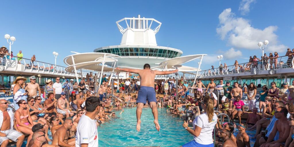 A man jumps into a crowded pool on a cruise ship, surrounded by cheering passengers on a sunny day.