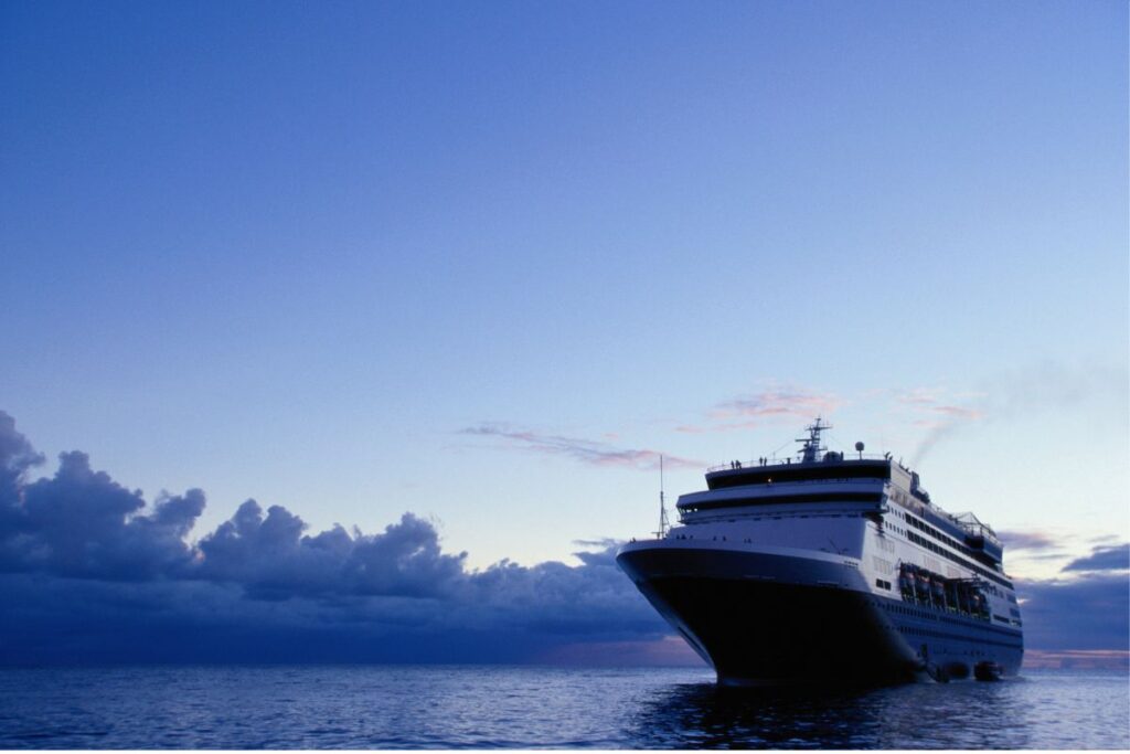 A cruise ship is seen sailing on calm waters under a vast, clear evening sky. The horizon is tinged with soft hues as the sun sets, casting a peaceful glow over the scene. The ship, captured from a low angle, appears majestic as it moves forward, with clouds in the distance adding to the tranquil atmosphere of the open sea.
