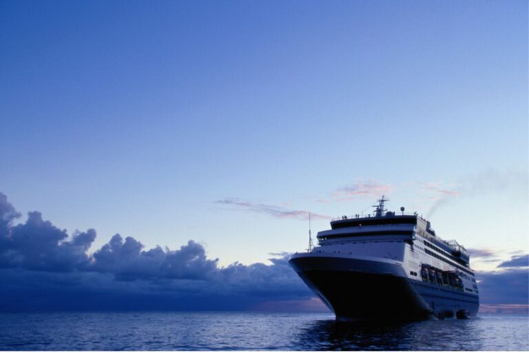 A cruise ship is seen sailing on calm waters under a vast, clear evening sky. The horizon is tinged with soft hues as the sun sets, casting a peaceful glow over the scene. The ship, captured from a low angle, appears majestic as it moves forward, with clouds in the distance adding to the tranquil atmosphere of the open sea.