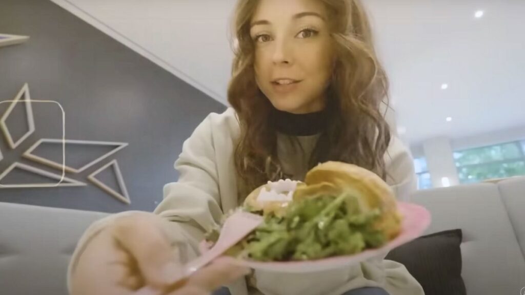 A young woman with long, wavy brown hair sits in a modern lounge area, holding out a pink plate with a croissant sandwich, a poached egg, and a fresh green salad. She wears a casual light-colored sweatshirt and has a friendly expression while engaging with the camera. The background features geometric wall art and a sleek, well-lit interior.