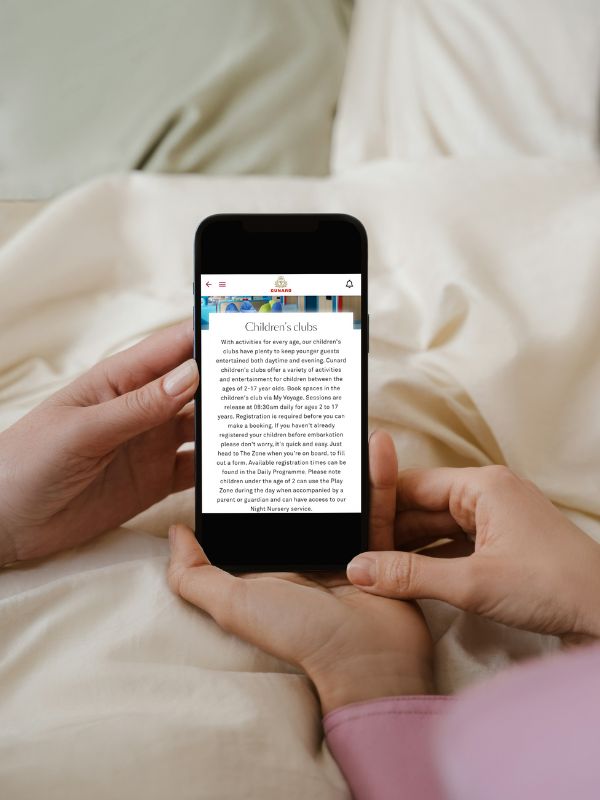 The image shows a person holding a smartphone displaying information about Cunard's Children's Club. The screen provides details about the various activities available for children aged 2 to 17 years, designed to keep younger guests entertained during their voyage. The text also covers registration processes and additional services such as the Play Zone and Night Nursery. The setting conveys a convenient and modern way for parents to access and manage information about children's programs on the cruise.