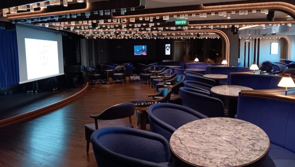 The image shows a sophisticated lounge or entertainment venue aboard Cunard's Queen Anne cruise ship. The room features plush, deep blue seating arranged around small marble-topped tables. Overhead, a modern lighting fixture with suspended square glass elements adds a chic touch. At the front, a stage with a piano and projection screen is set up, indicating this space is likely used for intimate performances, presentations, or talks. The ambiance is elegant and relaxed, offering a cozy yet stylish environment for guests to enjoy various onboard events.