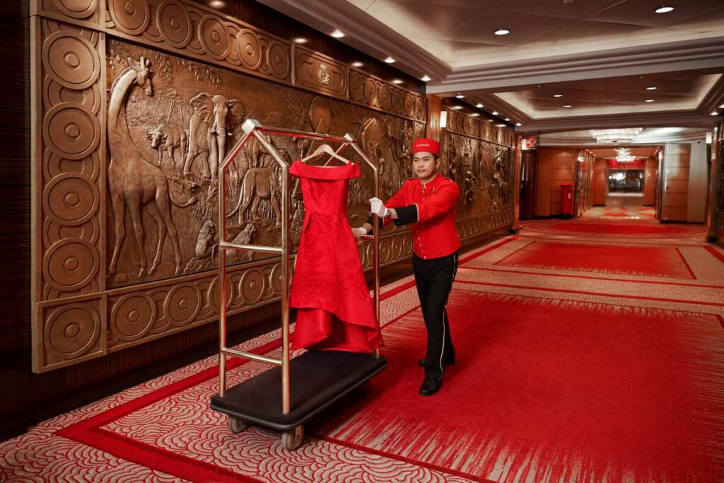 A bellboy in a red uniform is attentively positioning a red dress on a hanger on a portable clothes rack in the ornately decorated hallway of a Cunard cruise ship. The carpet is red with a patterned design, matching the aesthetic of the elaborate relief sculpture of animals on the wall.