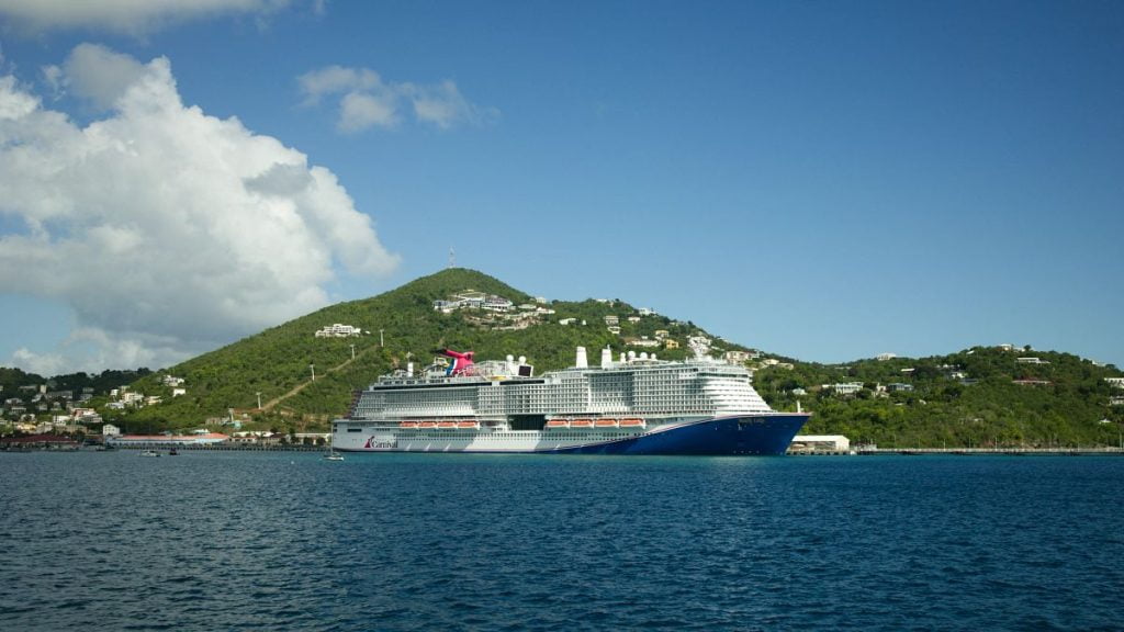 A Carnival cruise ship docked in a tropical port with lush green hills in the background and calm blue waters in the foreground under a partly cloudy sky.